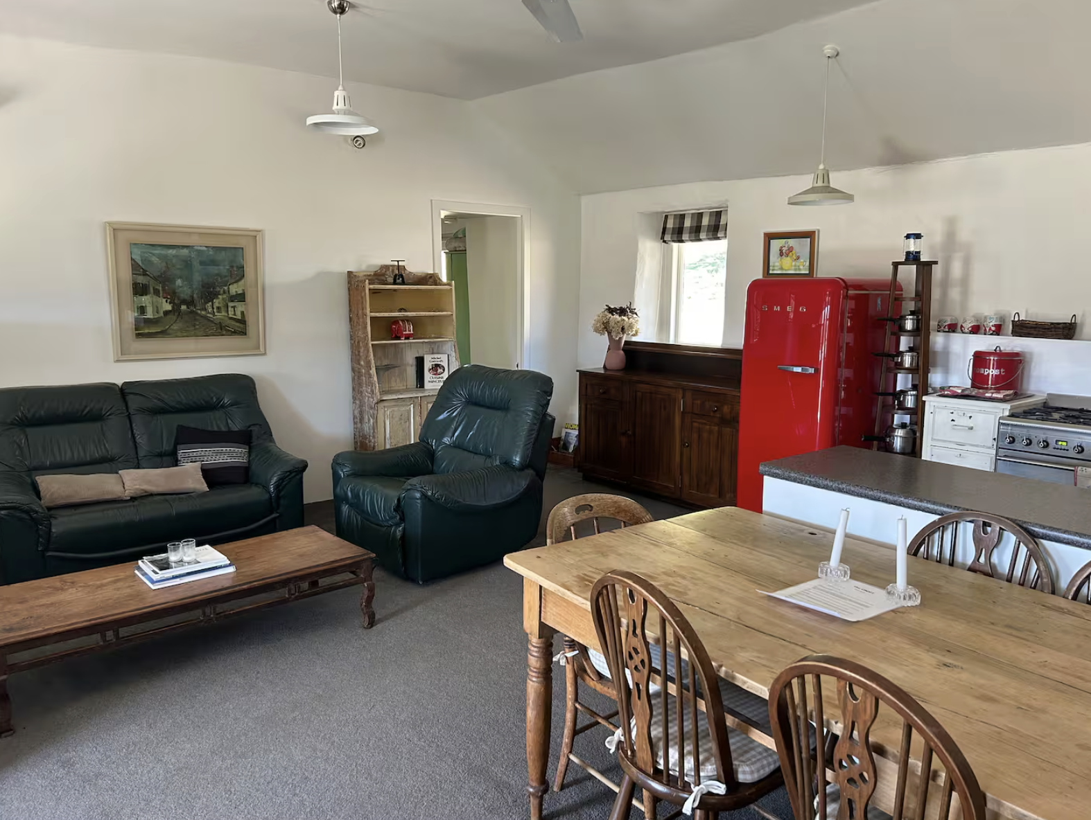 Living room with black leather sofa and armchair, wooden coffee table, dining table with chairs, vintage red SMEG refrigerator, wooden sideboard, shelves with dishes, window with checkered curtain, flowers on sideboard, painting on wall, ceiling fans, and kitchen appliances.