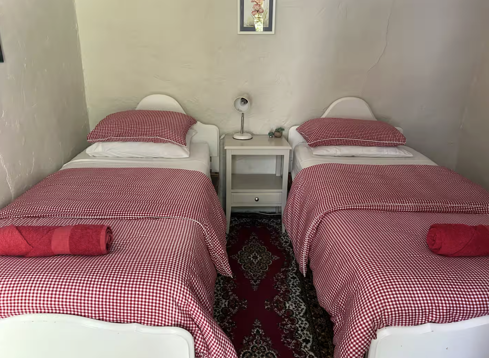 Two single beds with red and white checkered bedspreads and pillows, separated by a small white nightstand with a table lamp and decorative items on top, situated against a white wall with framed pictures, and a red and black patterned rug underneath.