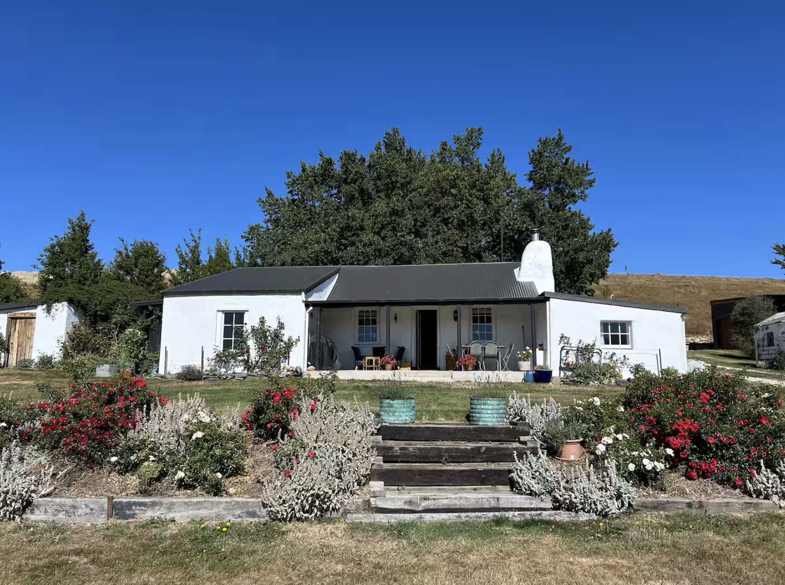 A white house with a dark metal roof, surrounded by colorful flowers and shrubs, with an outdoor seating area on the porch, set against a bright blue sky and a large tree in the background.