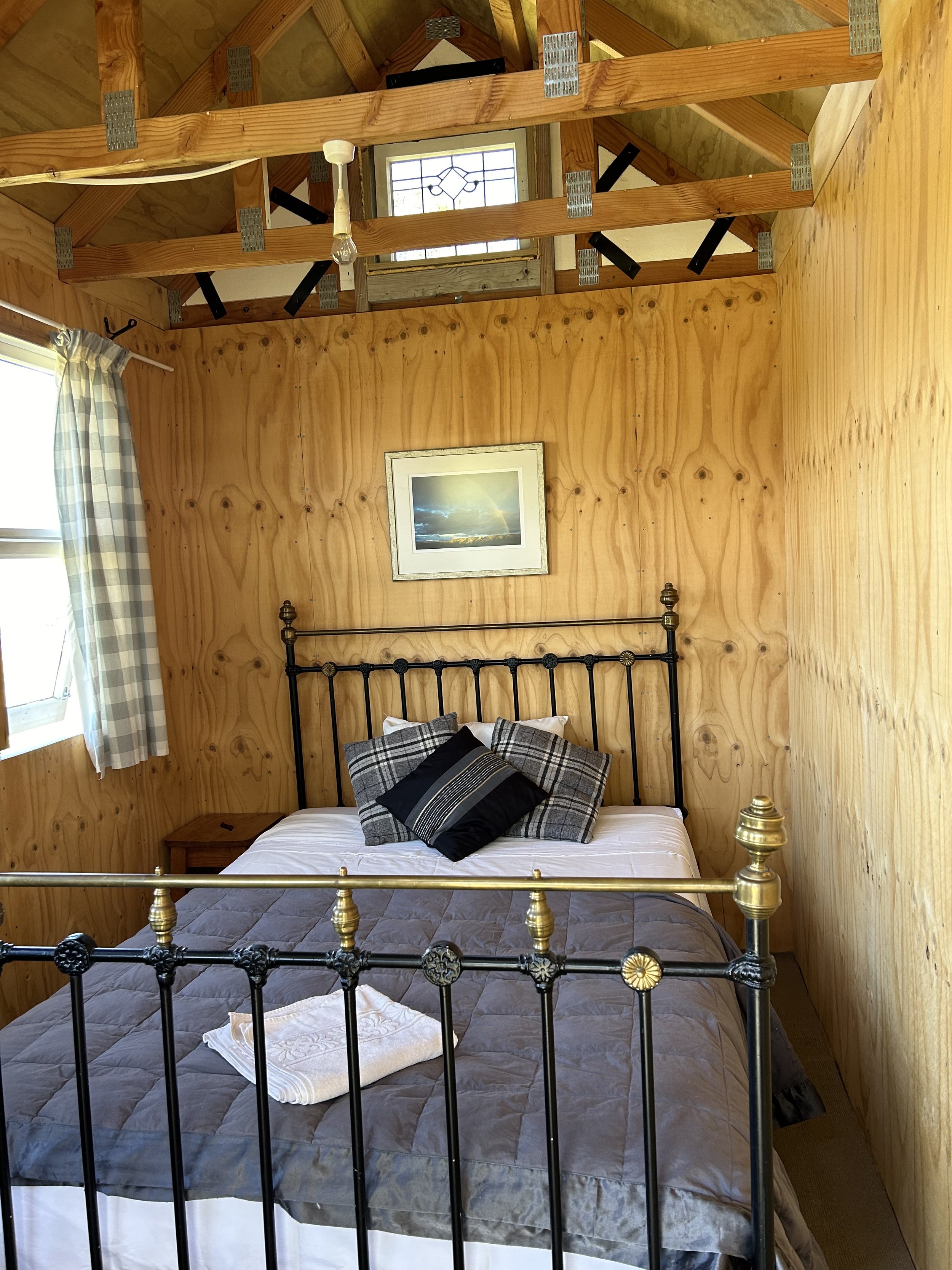 A cozy bedroom with wooden panel walls and ceiling framing. The bed has a black metal headboard and is made with white sheets, a gray quilt, and decorative pillows in plaid and black patterns. A nightstand is beside the bed, and a towel is folded on the foot of the bed. There is a window with checkered curtains on the left, allowing natural light into the room. A picture of a rainbow over clouds hangs above the bed.