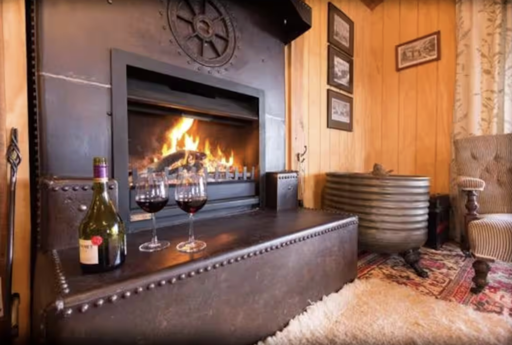 Living room with a lit fireplace, a bottle of red wine, and two glasses of wine on a wooden surface, beige armchair, wooden paneled walls, framed pictures, decorative carpet, and a curtain.