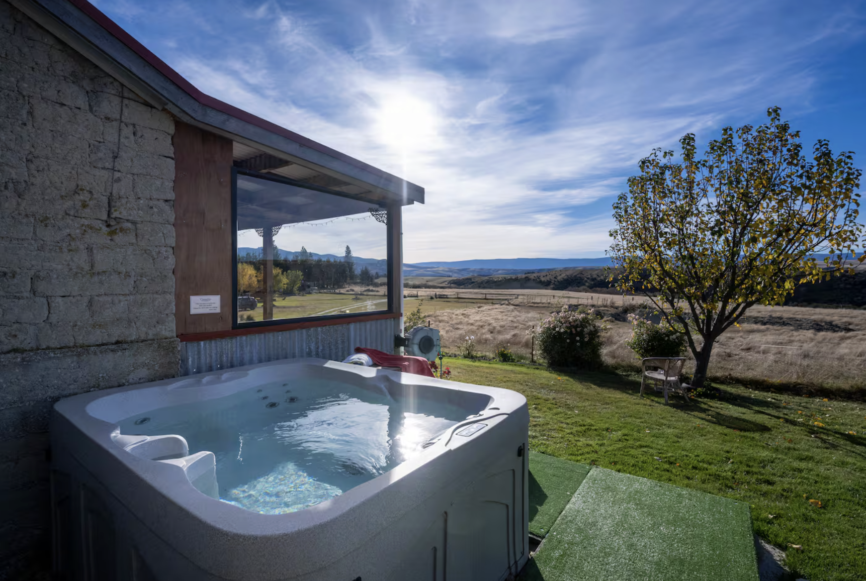 Outdoor hot tub on a patio, overlooking a rural landscape with fields, trees, and distant mountains during the daytime with clear blue skies.