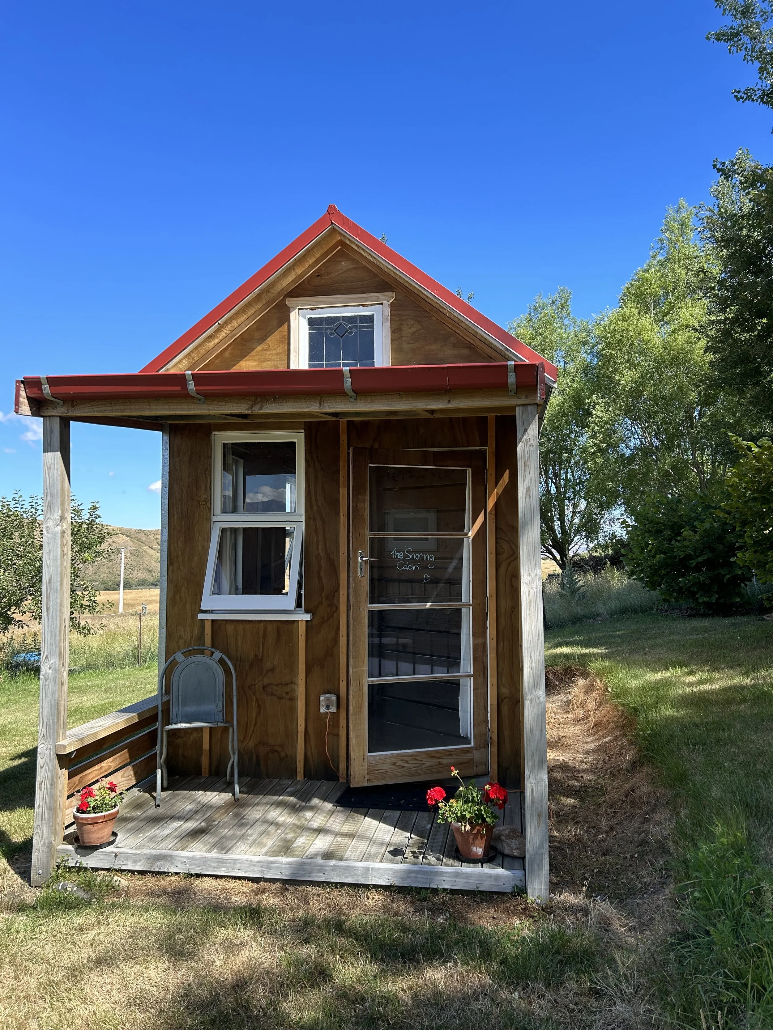 Small wooden cabin with a red metal roof, a front porch with potted flowers, a window, and a door with a sign that says "The Snoring Cabin". Surrounded by green grass and trees under a blue sky.