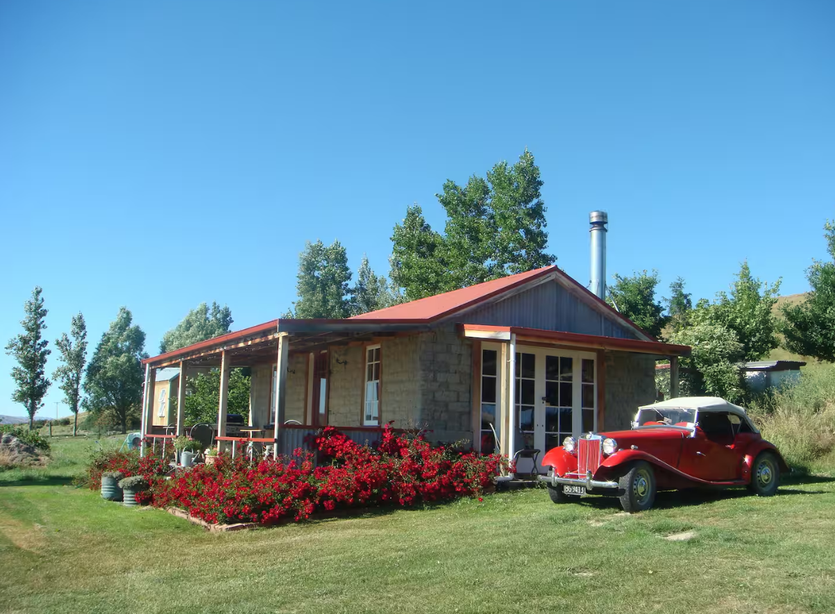 A small house with stone and wood exterior, surrounded by green grass and trees, with a vintage red car parked in front and red flowers in a flower bed.