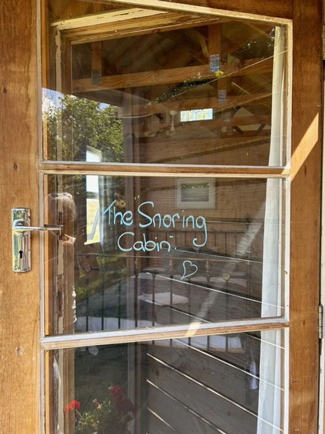 A wooden door with a glass windowpane that reads 'The Snoring Cabin' with a small heart. The interior of a rustic cabin is visible through the window, with wooden beams and walls.