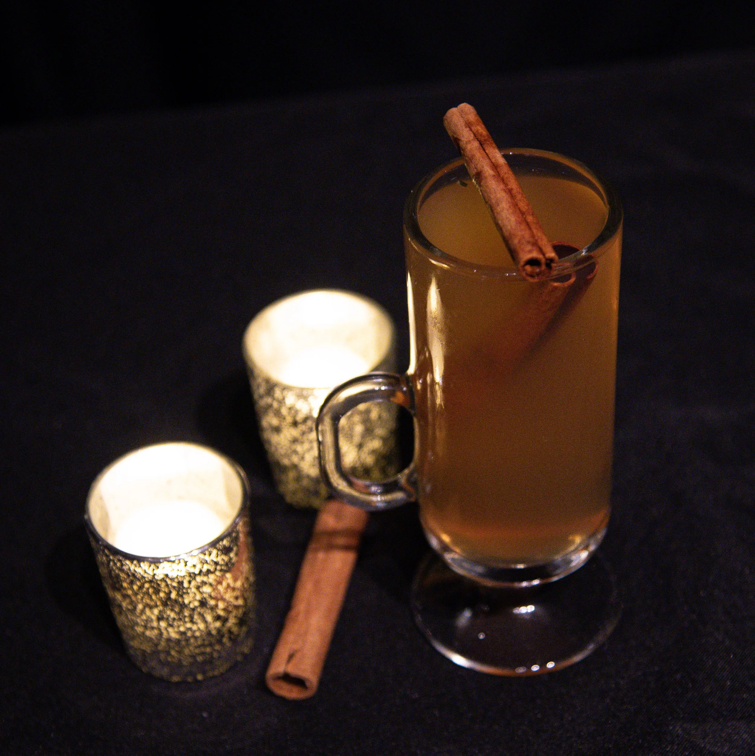 A tall glass of hot apple cider with a cinnamon stick garnish, accompanied by two lit decorative candles in gold holders on a dark surface.