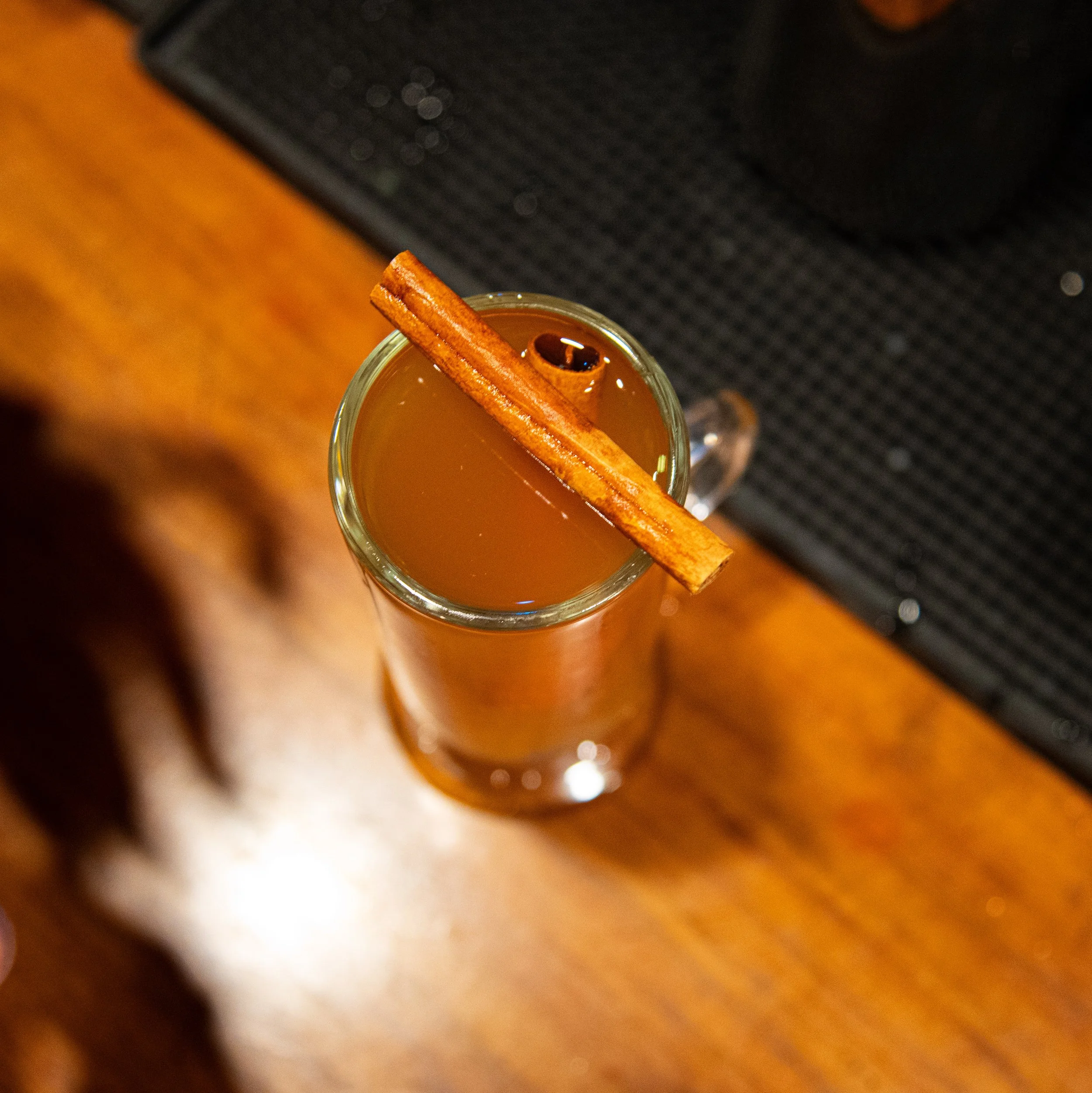 A hot beverage garnished with a cinnamon stick and a clove, served in a tall glass mug, placed on a wooden surface.