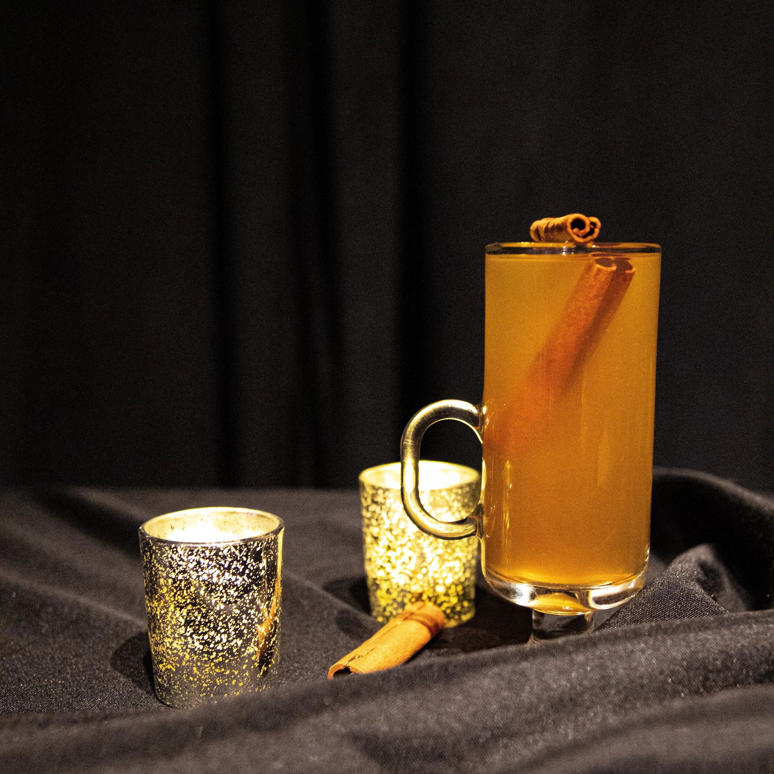 A tall glass with a handle filled with a warm, orange-colored beverage garnished with a cinnamon stick, alongside two small votive candles with perforated metal covers, and another cinnamon stick, all arranged on dark fabric against a dark background