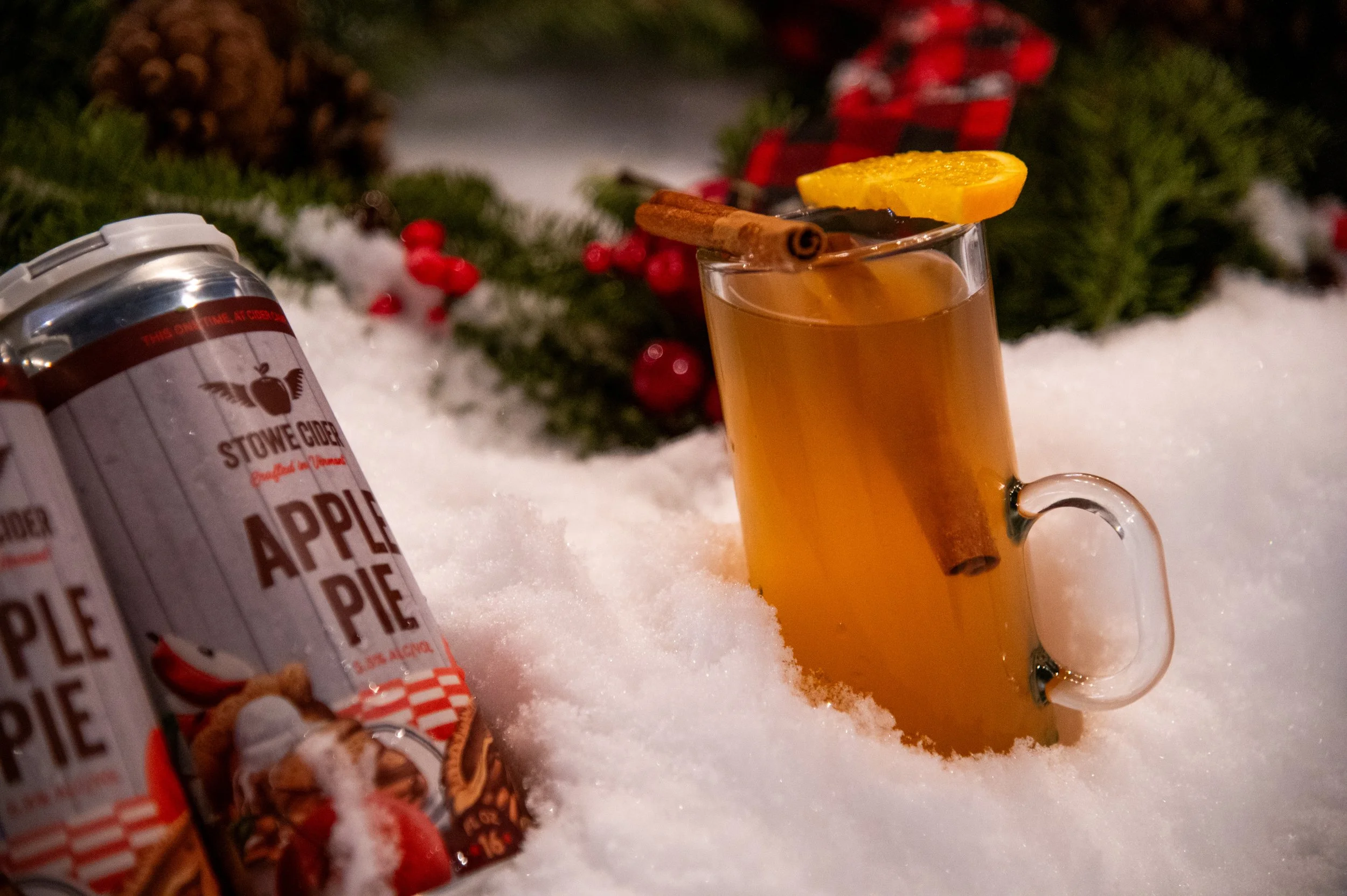 A hot beverage with a cinnamon stick and lemon slice garnishes in a glass mug, surrounded by snow, festive greenery, and red berries, with apple pie flavored drink cans nearby.