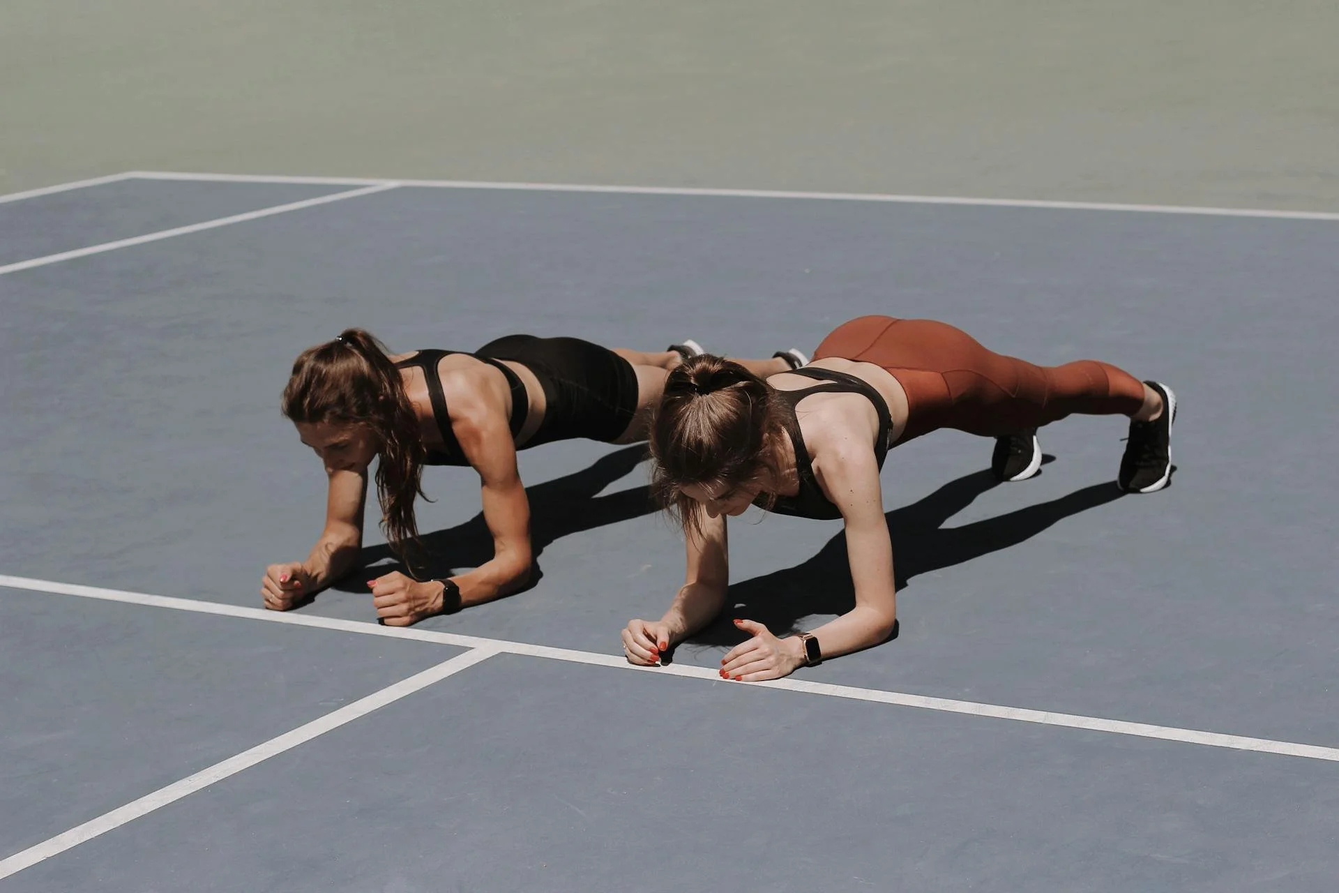 Two women are performing a plank exercise on a sports court, both in athletic wear and wearing fitness watches.