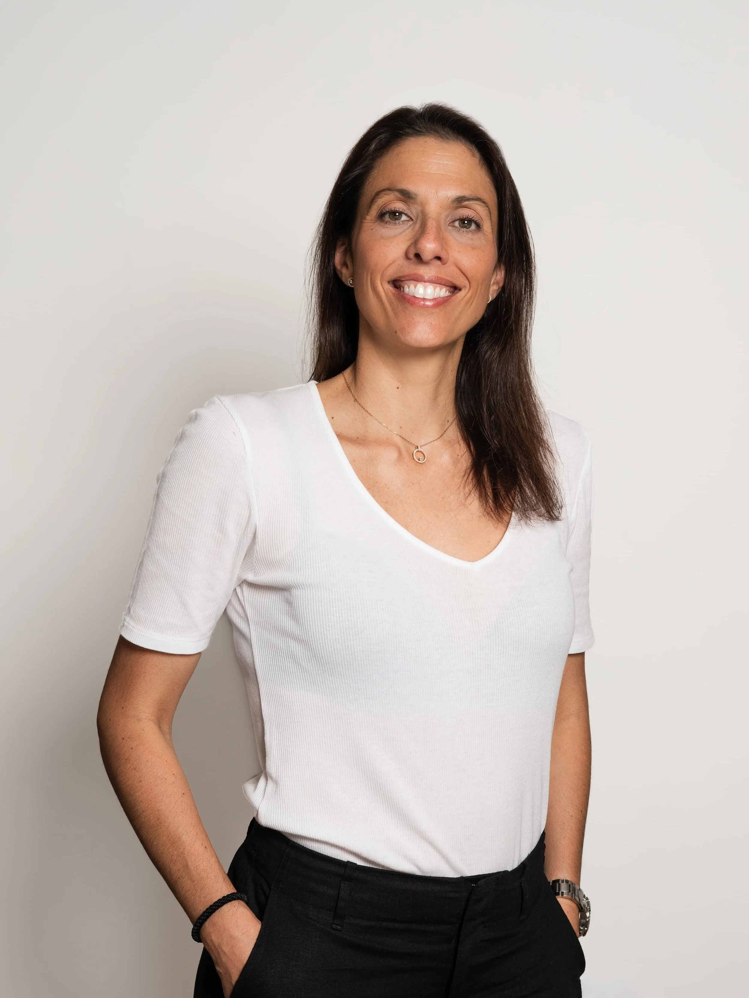A woman with long dark brown hair, wearing a white short-sleeve shirt, black pants, a gold necklace, and a watch, smiling and standing with her hands in her pockets against a plain white background.