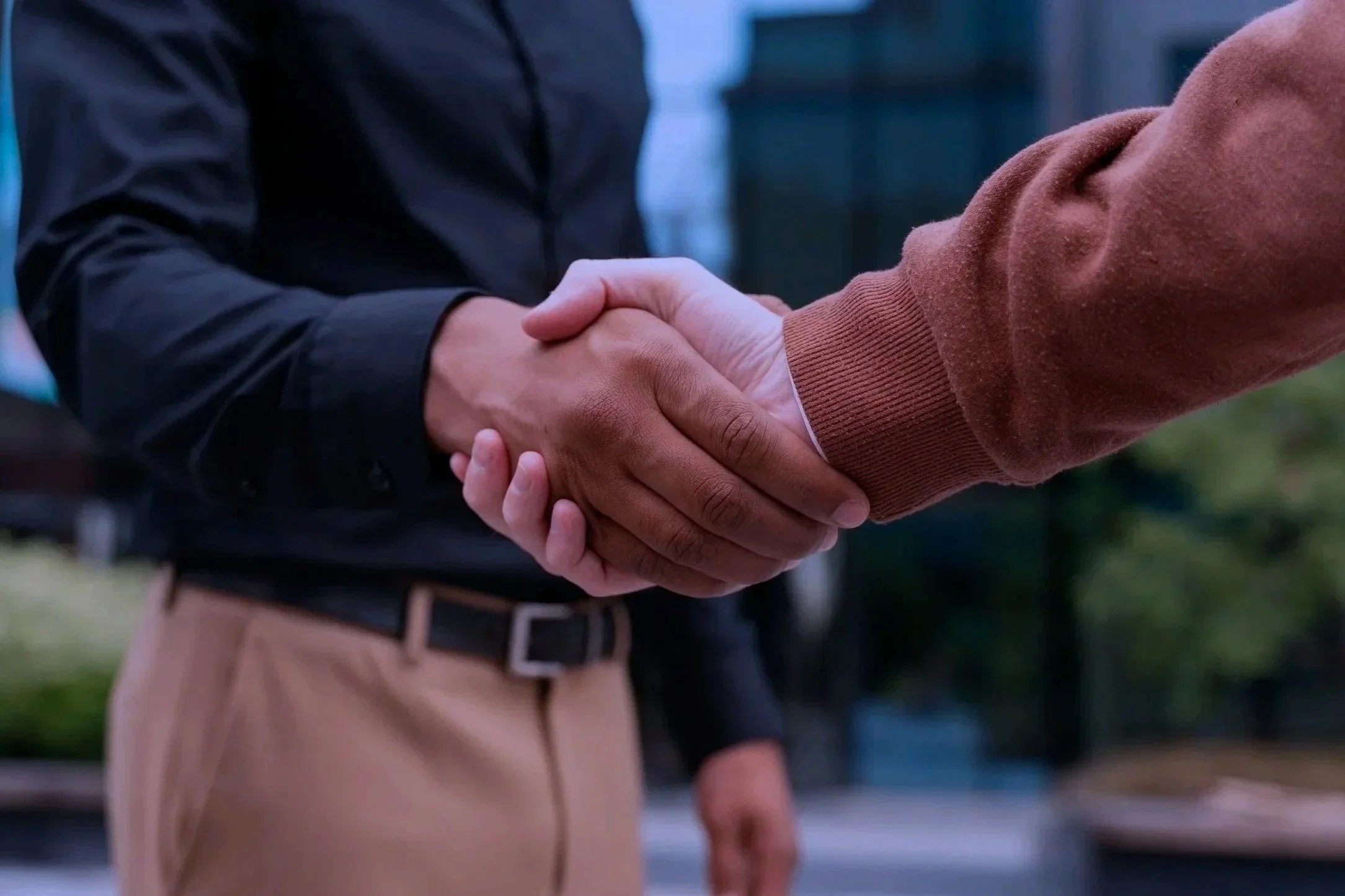 Two people shaking hands, one person wearing a brown long-sleeve shirt and the other wearing a black jacket, outdoors in a city setting.