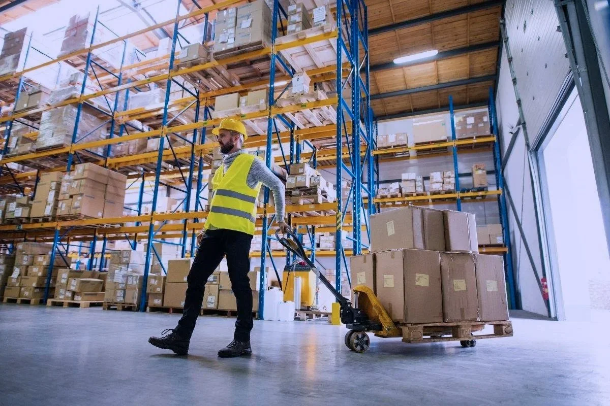 A warehouse worker in a yellow safety vest and helmet pushes a pallet jack loaded with boxes inside a warehouse with tall industrial shelves filled with boxes.