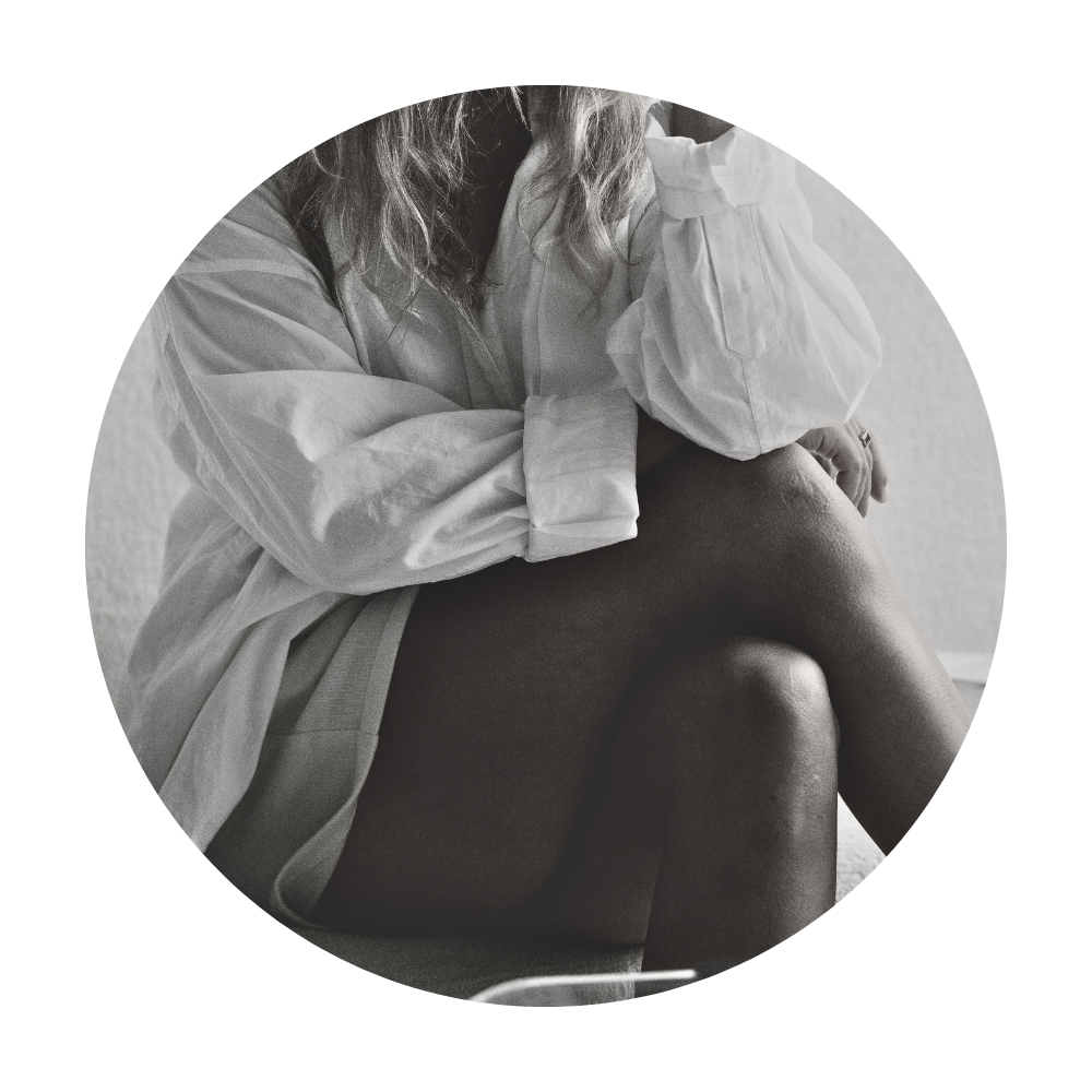 A woman sitting on a chair, wearing an oversized shirt, with her legs crossed. The photo is in black and white.