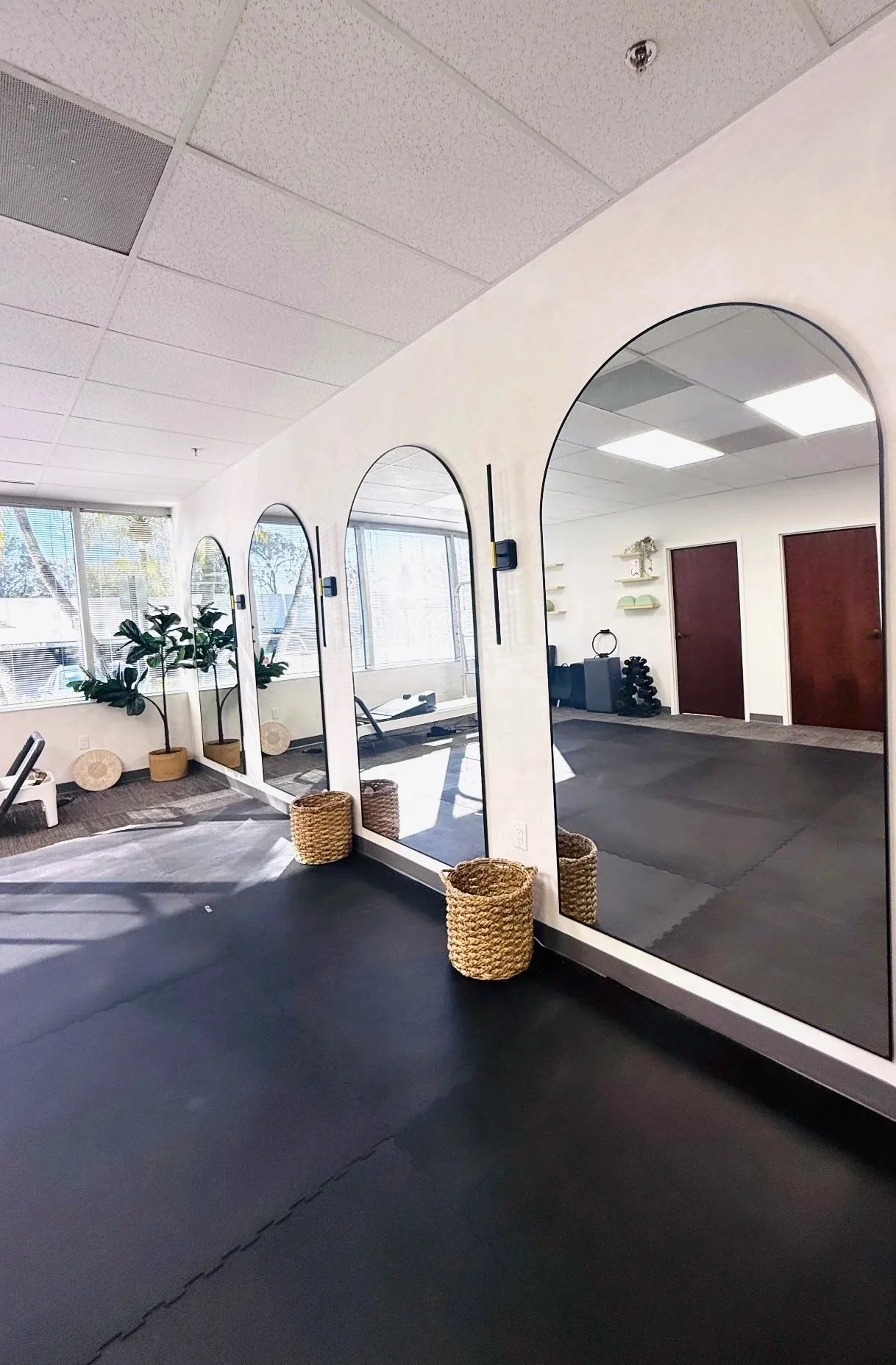 A fitness studio with three large arched mirrors on the wall, potted plants near windows, and wicker baskets on the floor, with workout equipment visible in the reflection.