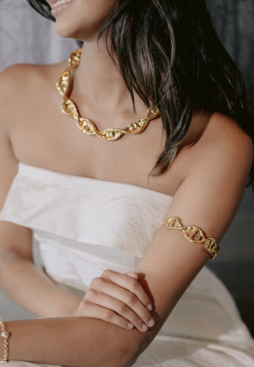 A woman wearing gold jewelry, including a twisted necklace and arm bracelet, against a plain background, dressed in a white strapless dress.