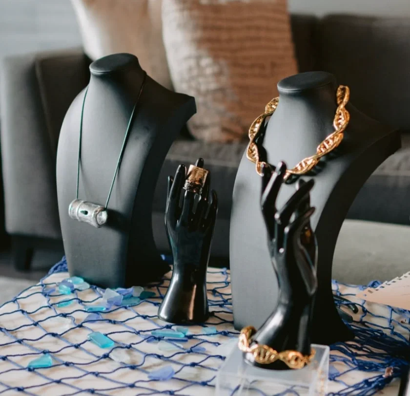 Jewelry display with necklaces and bracelets on black busts and hand-shaped holders, set on a table with blue and white netting and scattered blue stones.