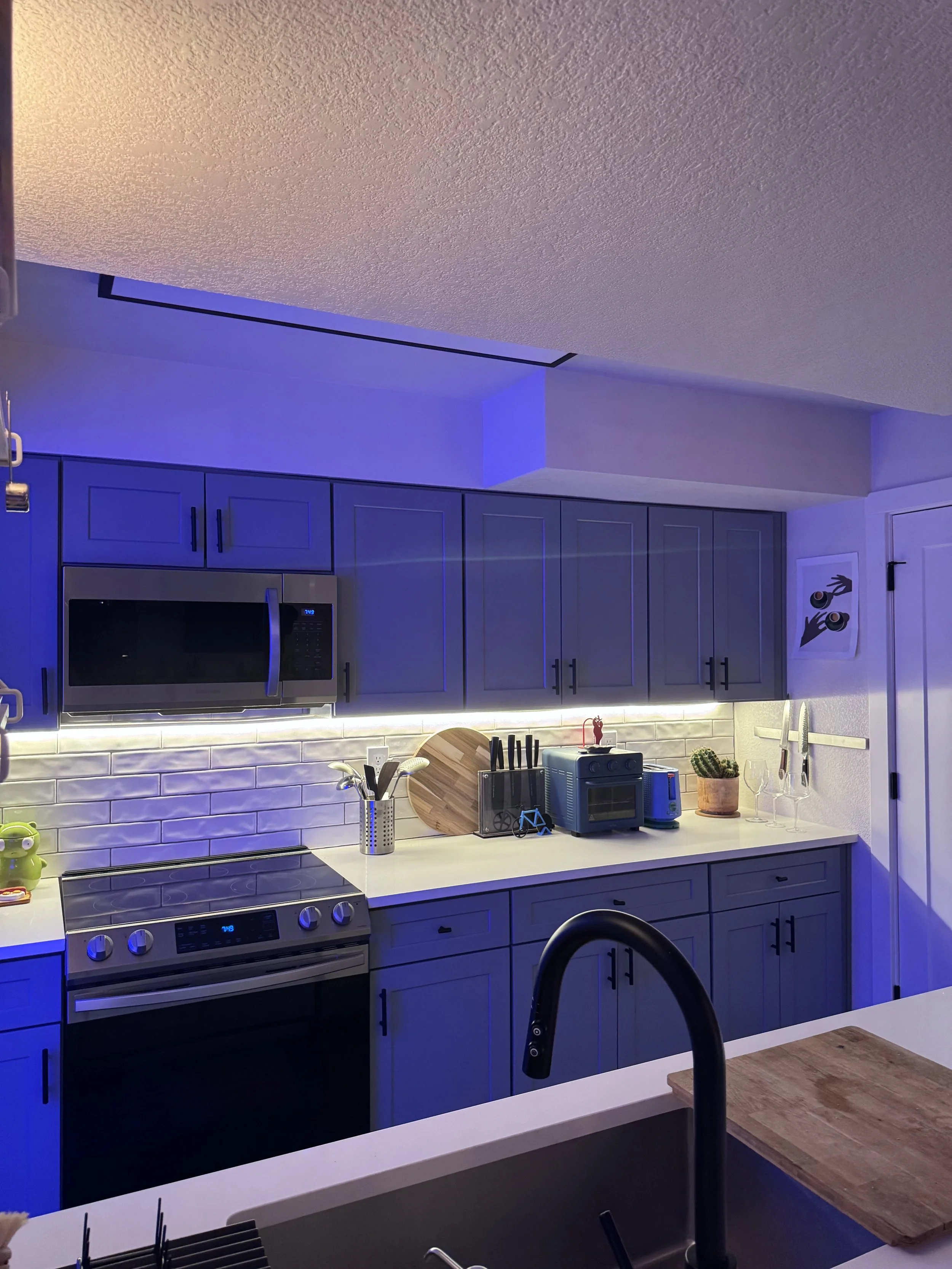 A modern kitchen with blue cabinets, white countertops, and a white subway tile backsplash illuminated by under-cabinet lighting. There is a stove, microwave, toaster, cutting board, utensils, potted cactus, wine glasses, and various small appliances on the counter.