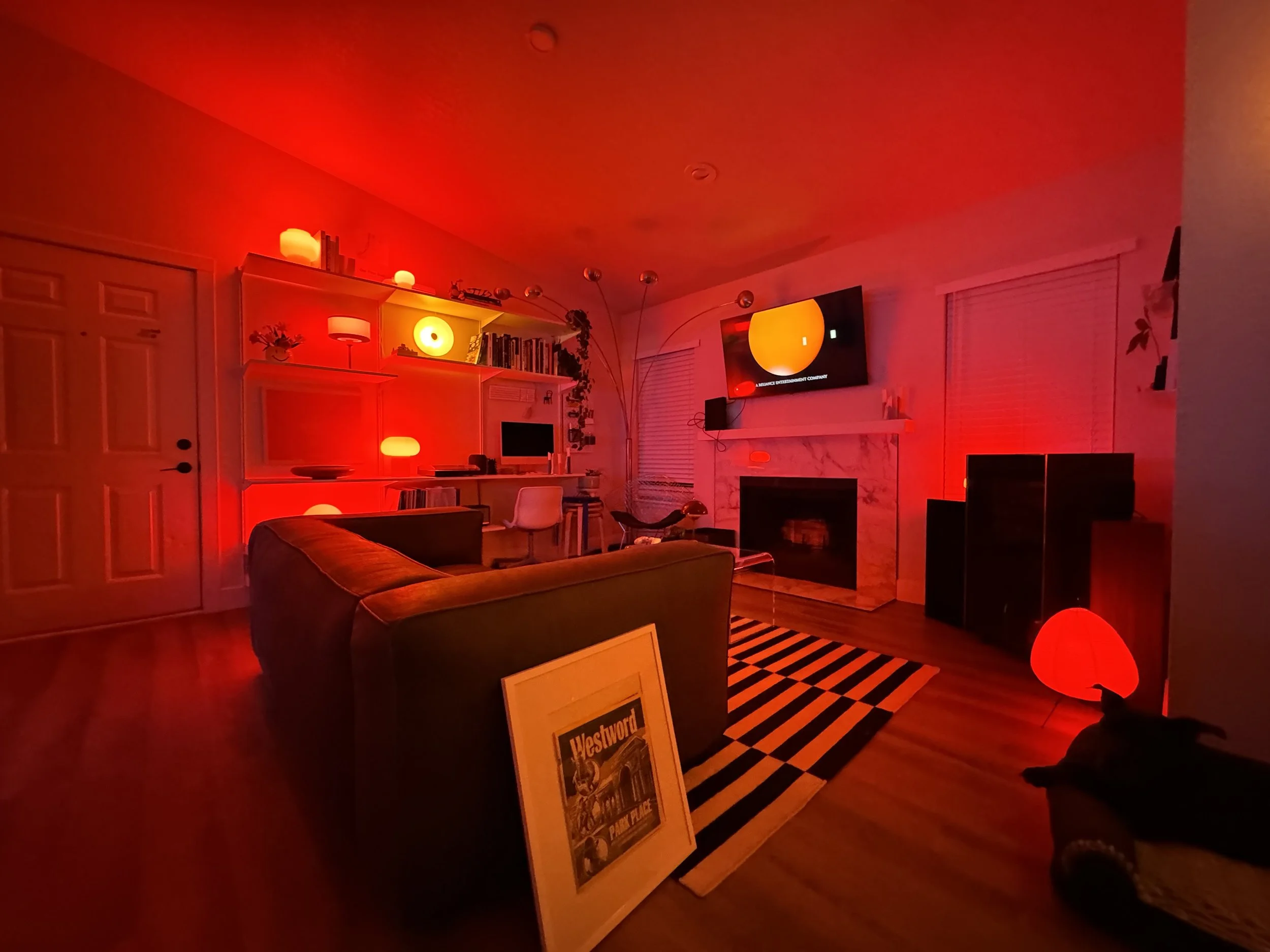 Living room illuminated with red lighting. Features a dark sofa, a black and white striped rug, a fireplace, a wall-mounted TV, bookshelves, and various decorative items.