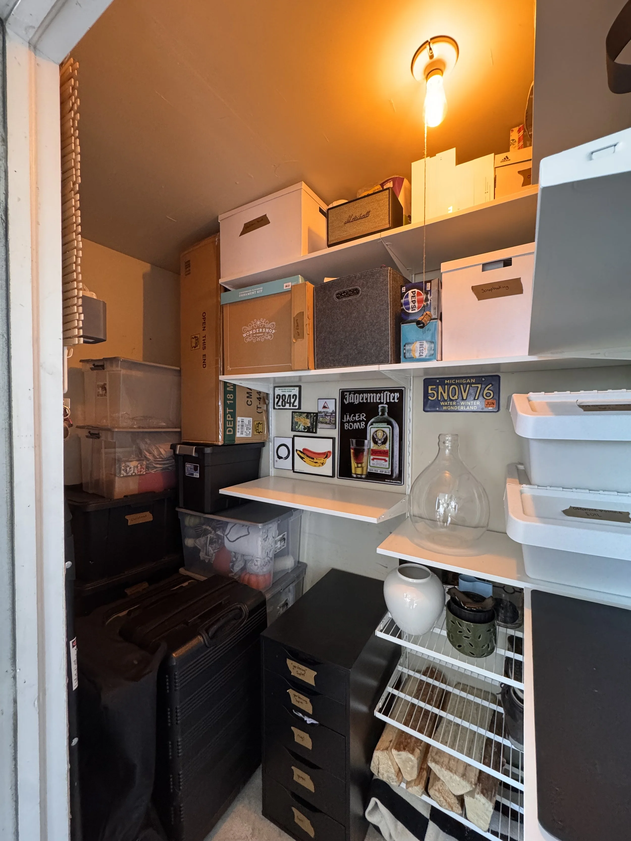 A small storage closet with shelves filled with boxes, containers, decorations, and miscellaneous items, illuminated by an orange ceiling light.