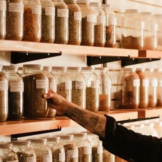 Shelves of large jars of herbs shown at an angle with an arm holding one of the jars.