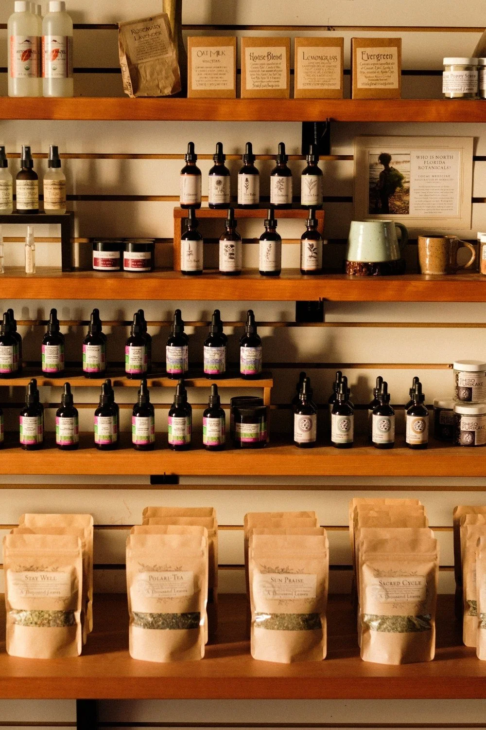 Shelves on a wall displayed with tincture dropper bottles, brown bags of loose tea, spray bottles, and glass pots with herbal balms.
