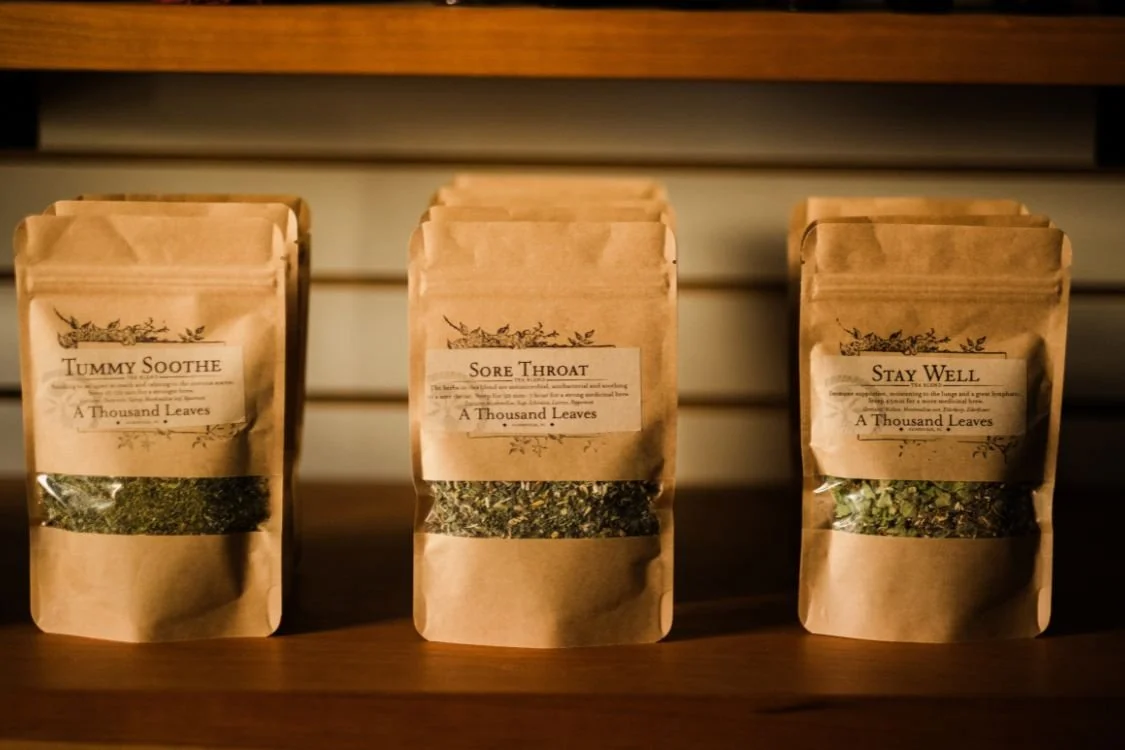 Three brown paper packets labeled 'Tummy Soothe,' 'Sore Throat,' and 'Stay Well' with dried herbs inside, placed on a wooden surface.