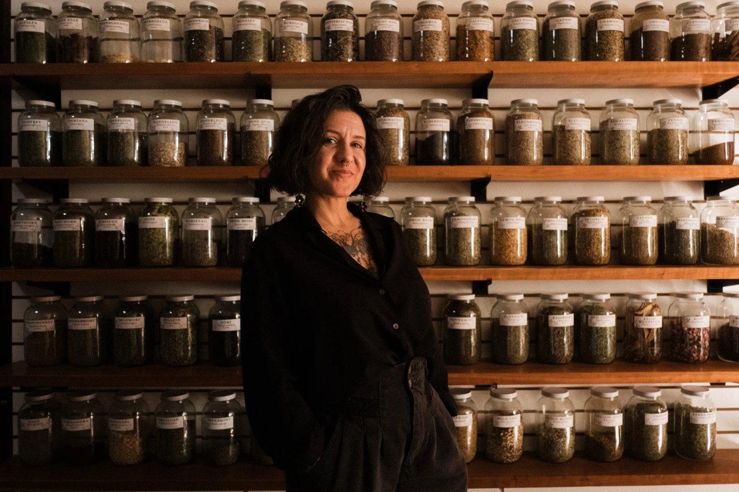 A woman with short dark hair and wearing all black standing in front of a wall of shelves lined with large jars filled with herbs, she is smiling at the camera.