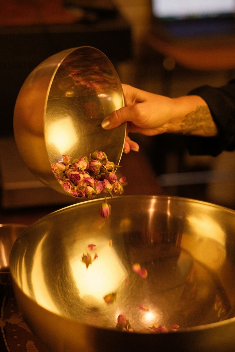 A hand covered in tattoos pours loose rose buds out of a golden bowl into a larger golden bowl.
