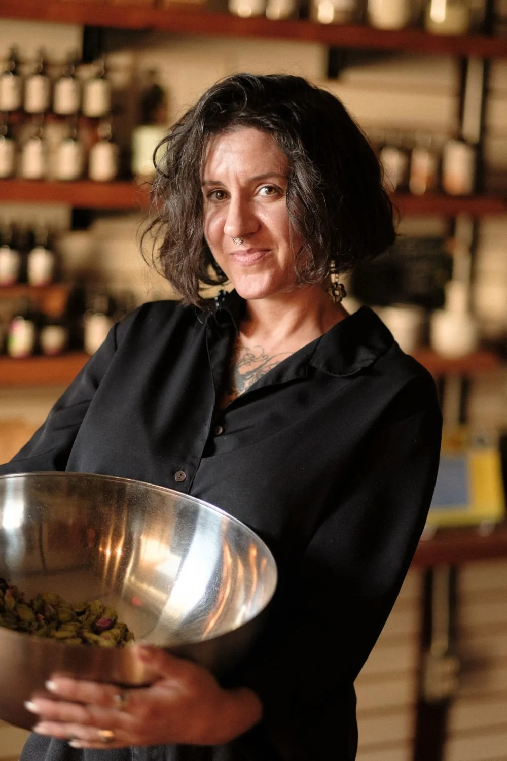 A woman with short dark hair wearing a black blouse smiling at the camera and holding a large silver bowl with herbs