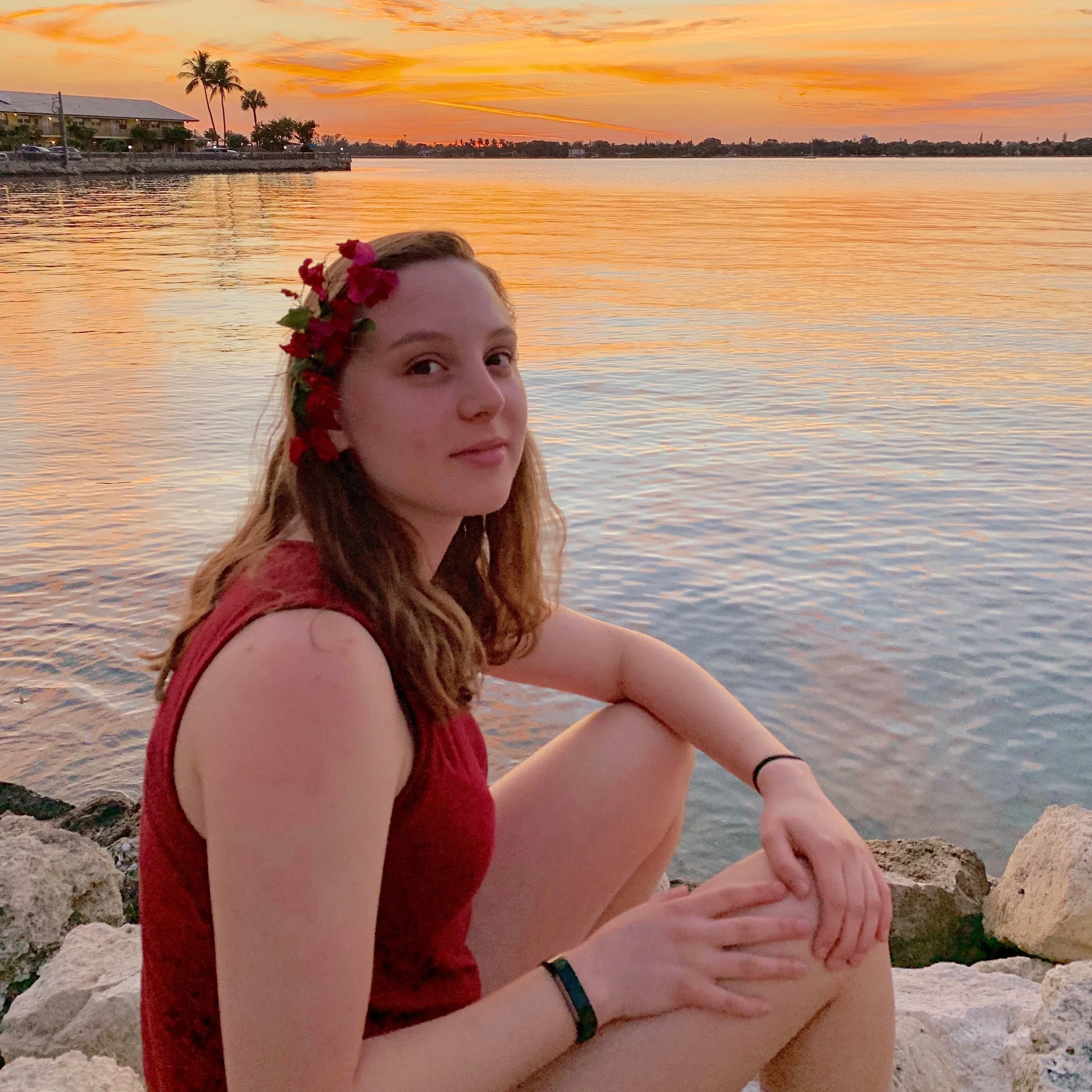 A woman with long brownish hair with flowers sitting in front of water at sunset and smiling at the camera.