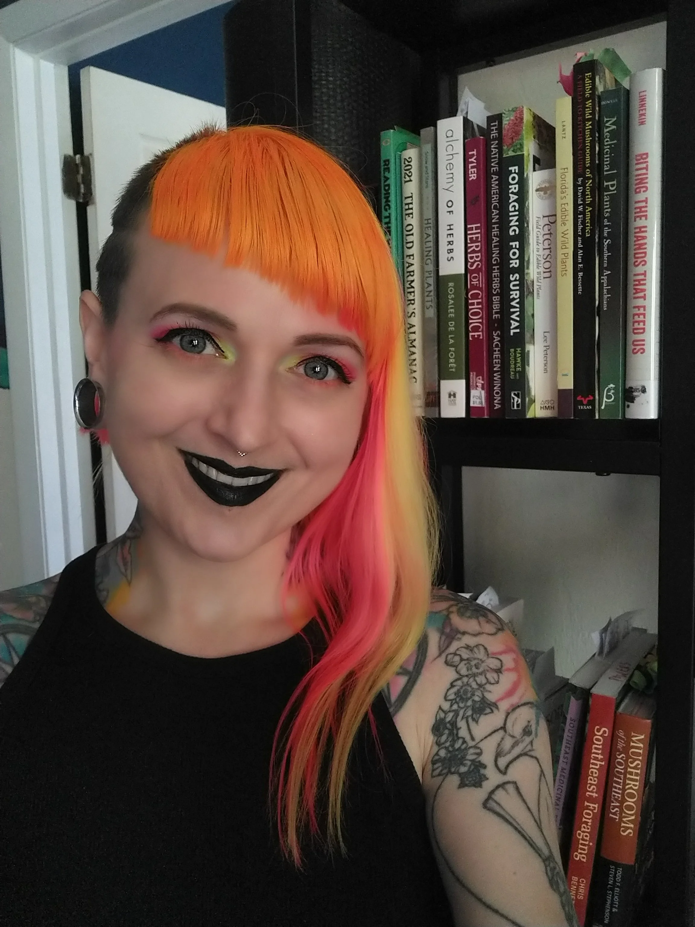 A portrait of a woman with orange and pink hair and dark lip color looking at and smiling at the camera.