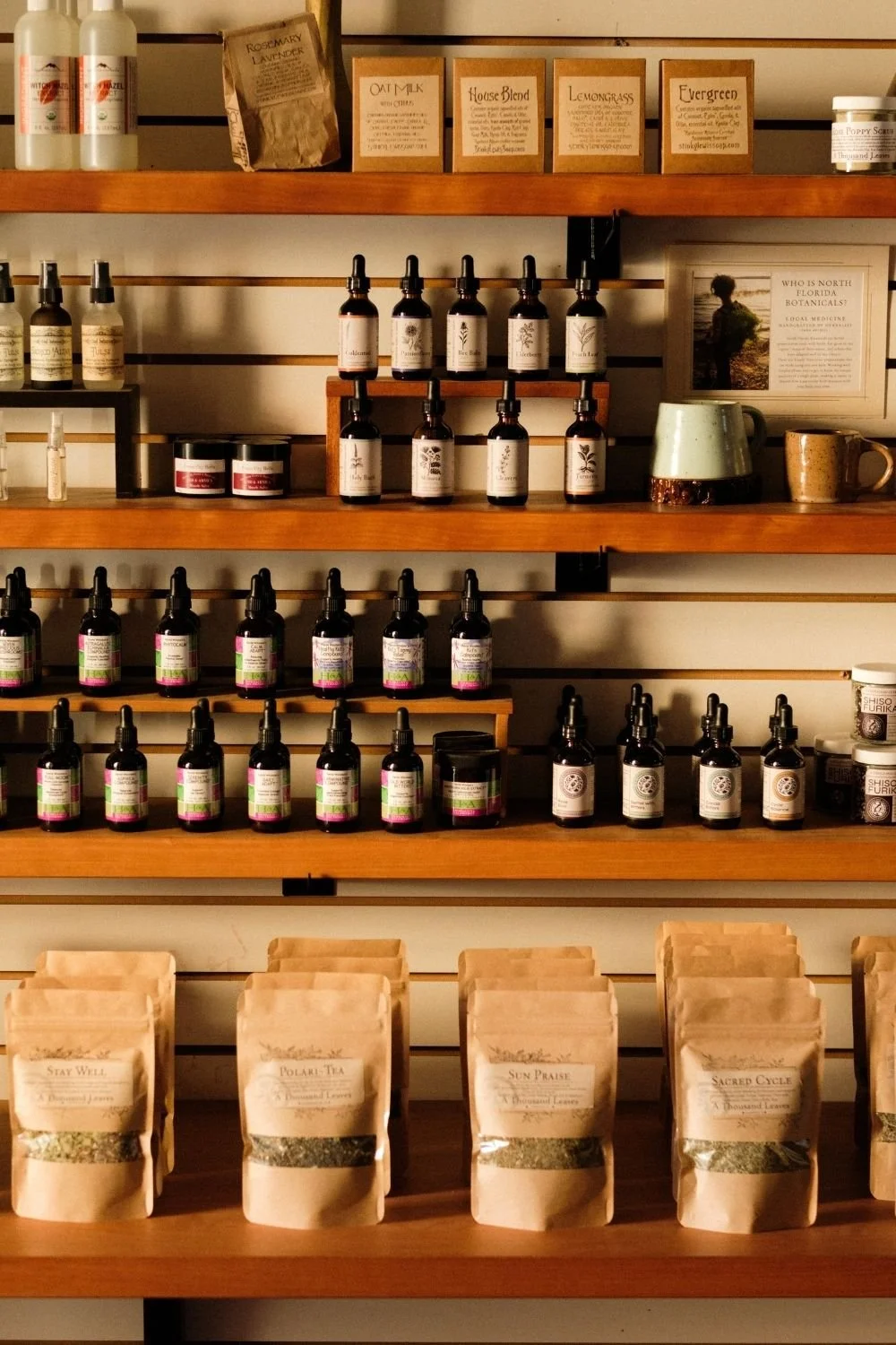 Shelves on a wall displayed with tincture dropper bottles, brown bags of loose tea, spray bottles, and glass pots with herbal balms.