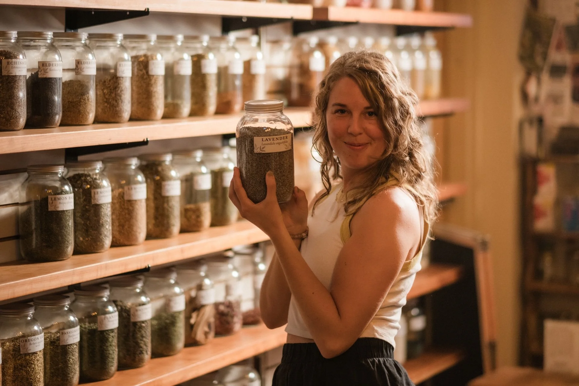 A woman with short light brown hair and a necklace with a crescent moon pendant, smiling in front of shelves filled with glass jars of herbs and spices labeled with their names.