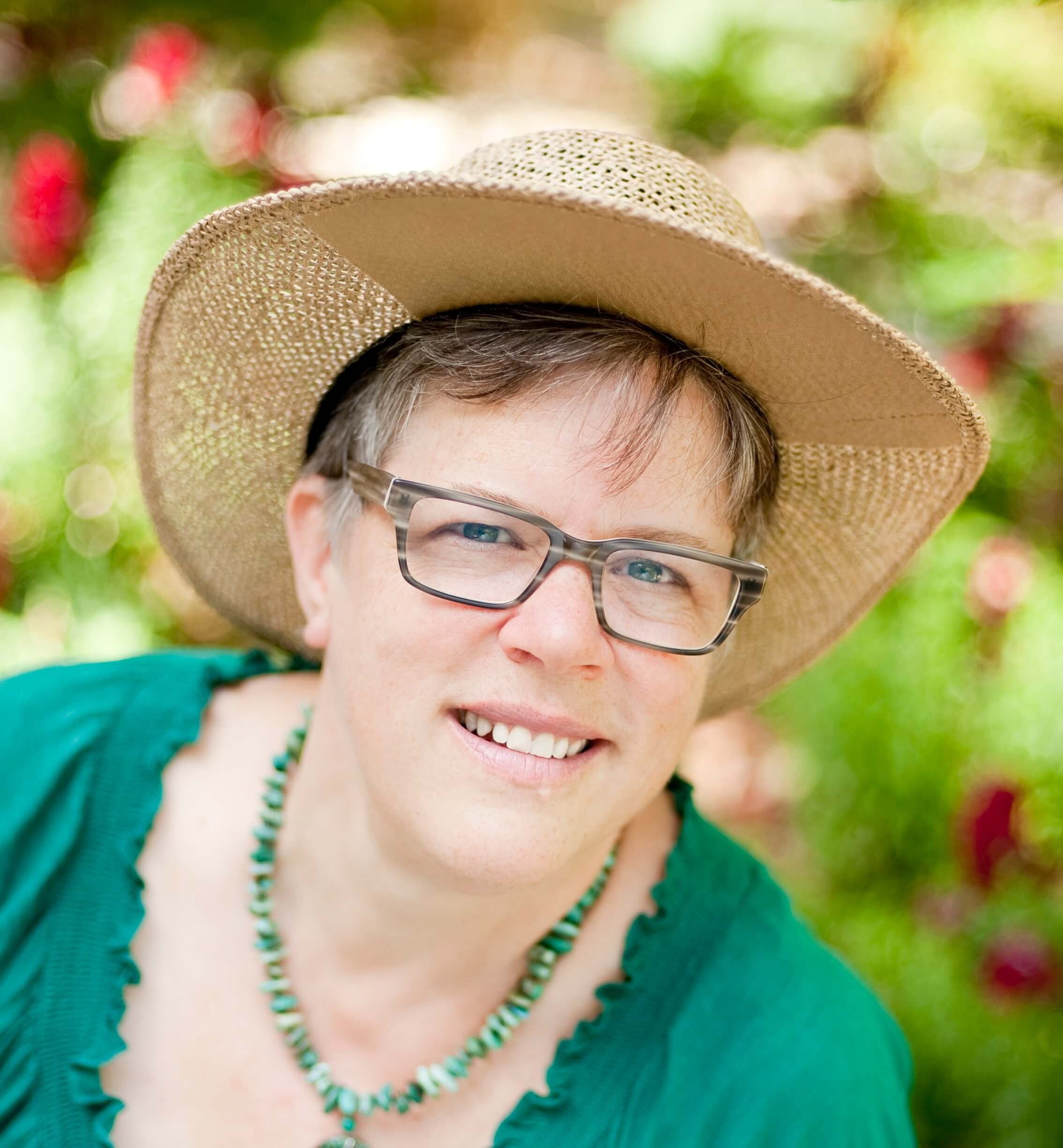 A smiling woman with short hair wearing glasses, a wide-brimmed straw hat, a green shirt, and a turquoise beaded necklace, outdoors with green and pink blurred foliage in the background.