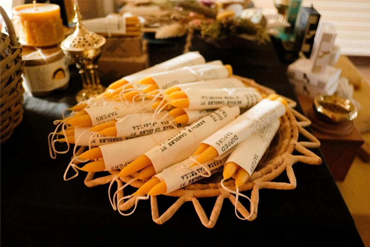 A woven basket filled with pairs of yellow beeswax candles wrapped in paper and labeled Dipped Tapers.