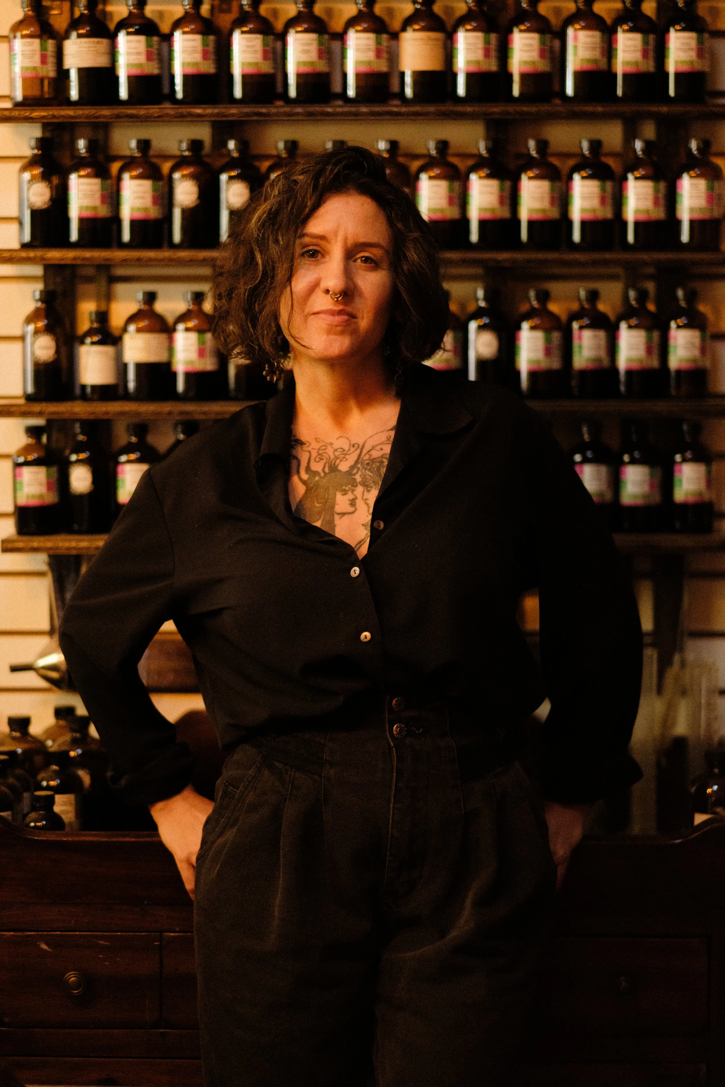 A woman with short, curly hair, wearing a black shirt and high-waisted black pants, standing in front of wooden shelves filled with bottles in a store or apothecary.