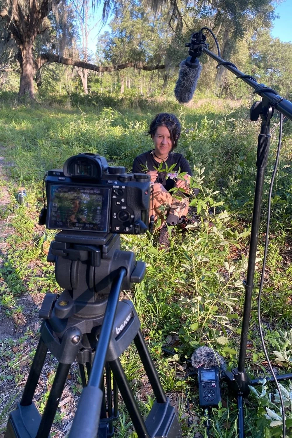 A digital camera on a tripod records a woman with dark hair and dark clothes sitting on the ground surrounded by plants and smiling at the camera.