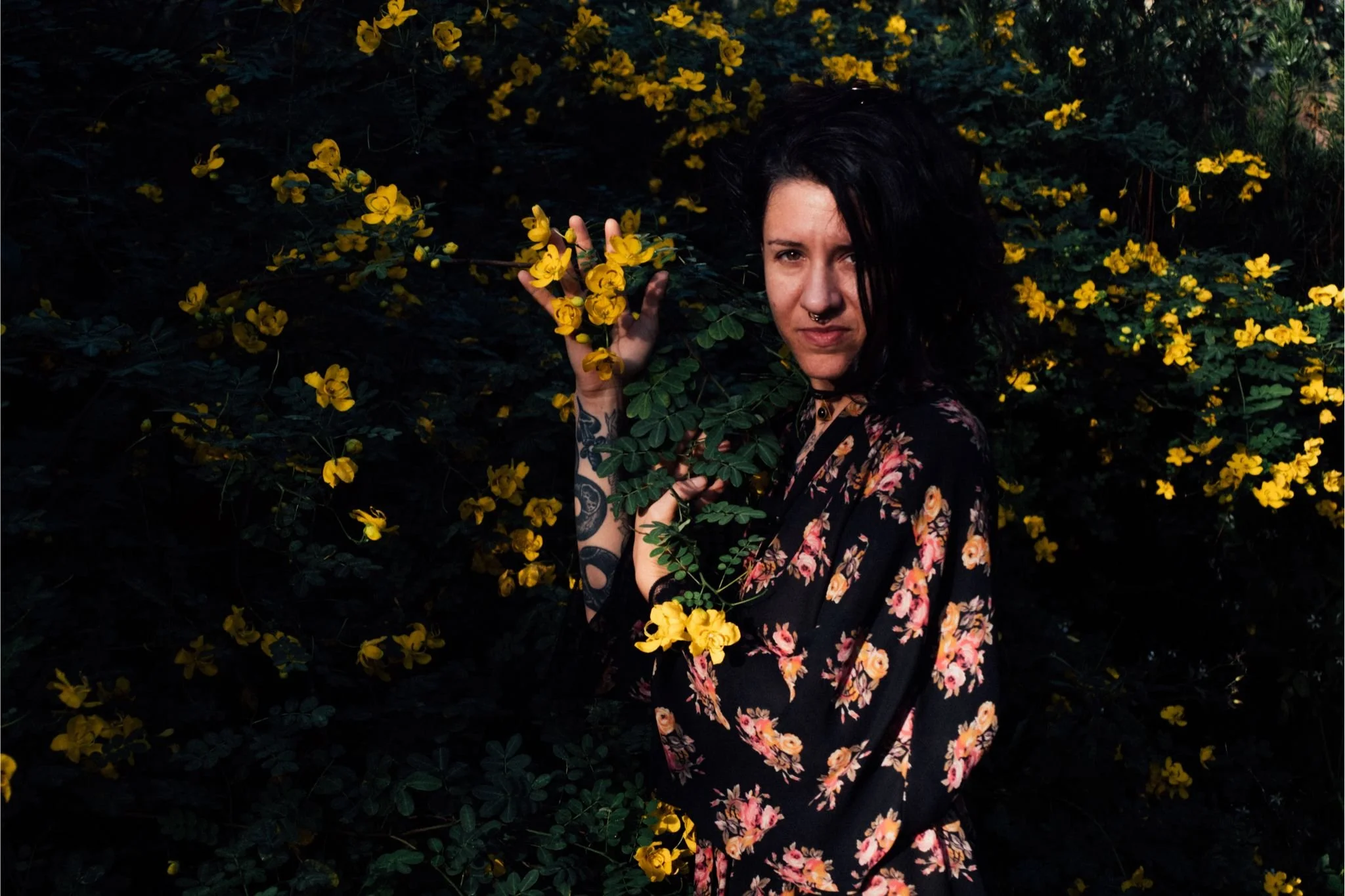 Woman with dark hair and tattoos on her arm wearing a black floral dress, standing among dark green foliage and yellow flowers