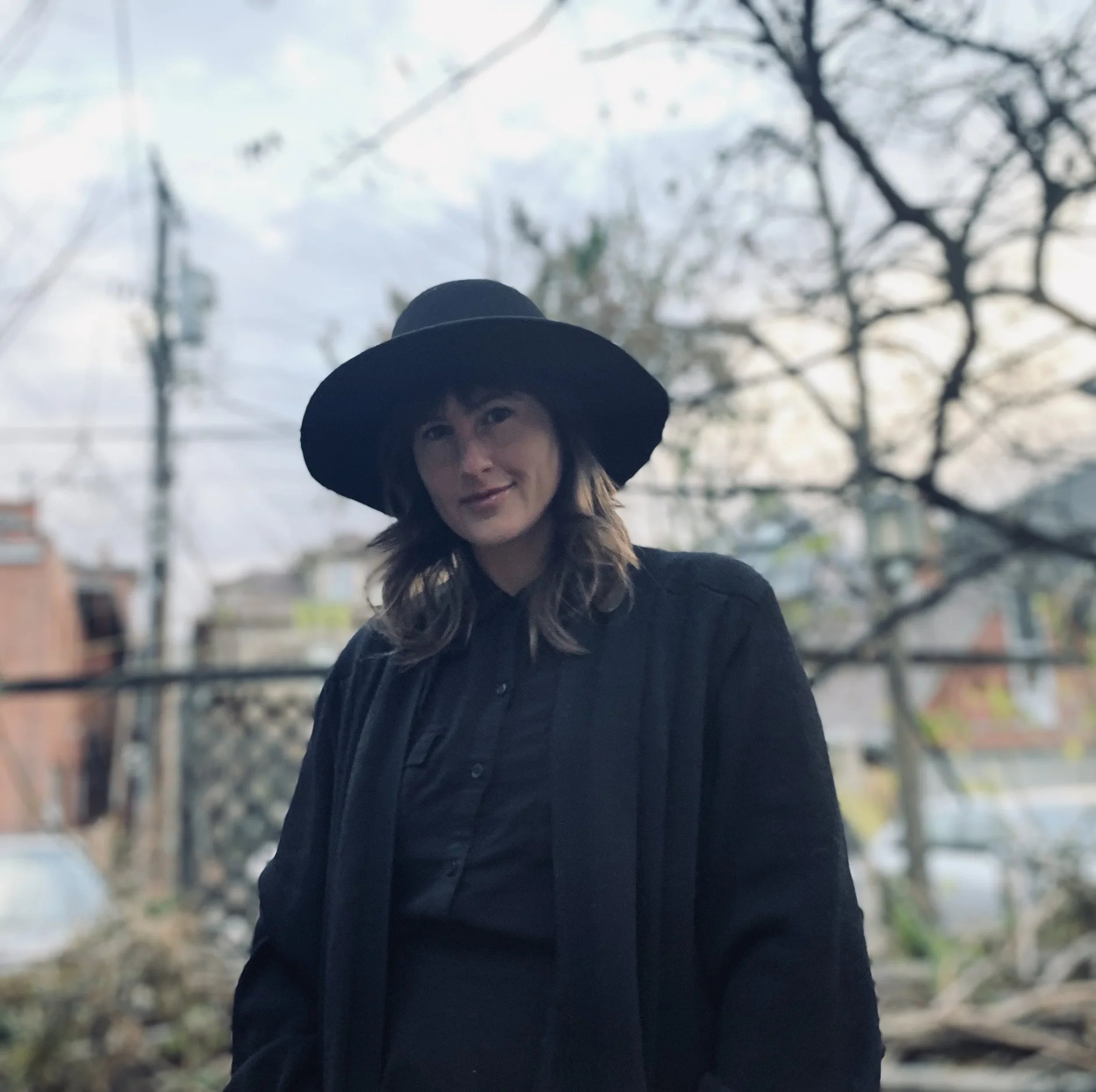 A woman with wavy brown hair wearing a large black hat and black clothing stands outdoors against a blurred background of leafless trees and buildings during daytime.