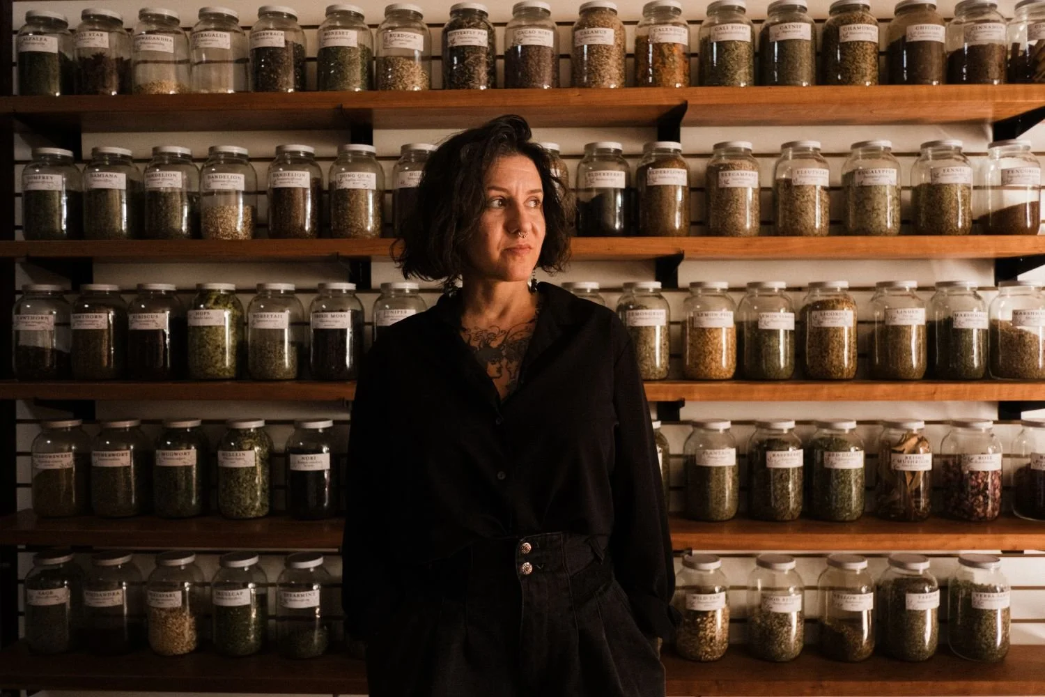 A woman with short dark hair and wearing all black looks off to the side while standing in front of a wall lined with shelves of jars filled with herbs.