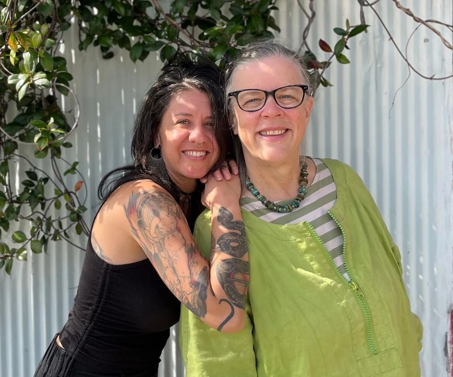 Two women smiling at the camera. One has long dark hair and wearing a black sleeveless shirt is leaning on the shoulder of the other, older woman with white and gray hair and glasses wearing a bright green jacket.