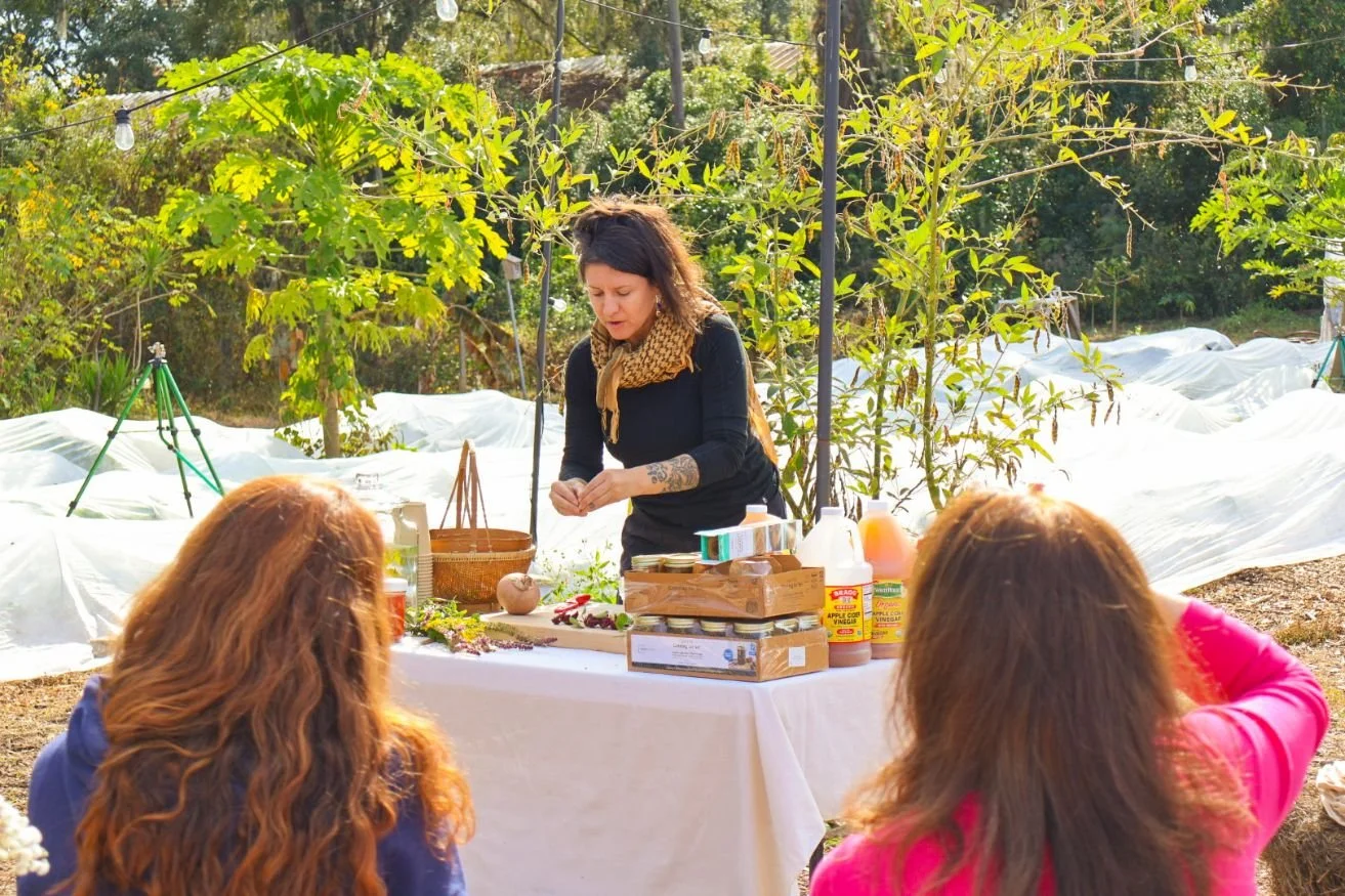 A woman standing  and talking behind a table covered with jars, food, and bottles of vinegar