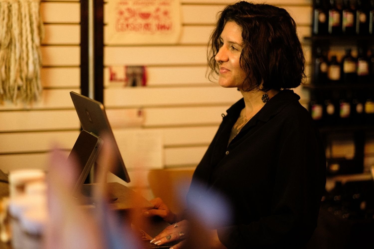 A woman with short dark hair wearing a black blouse standing at a checkout counter looking over the counter and smiling.