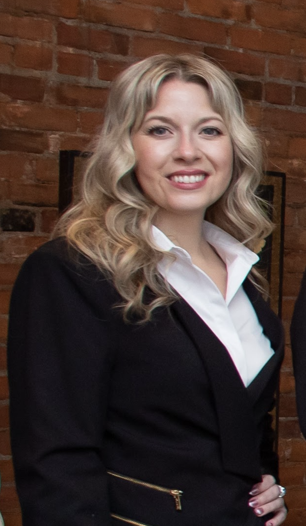 A woman with blonde wavy hair smiling, dressed in a white shirt and black blazer, standing against a brick wall.