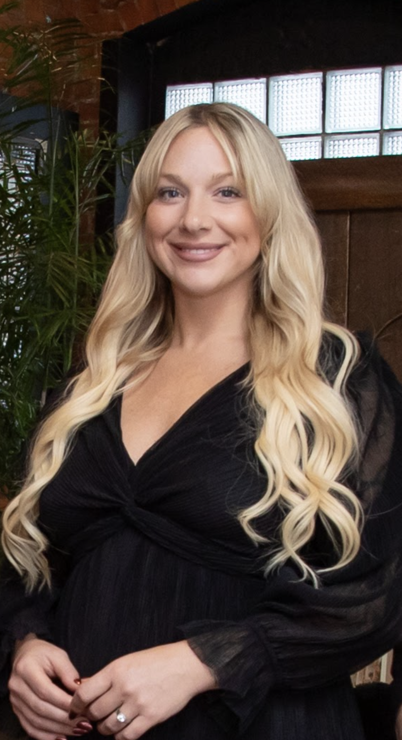 A smiling woman with long blonde wavy hair wearing a black dress standing indoors, with green plants and a window in the background.
