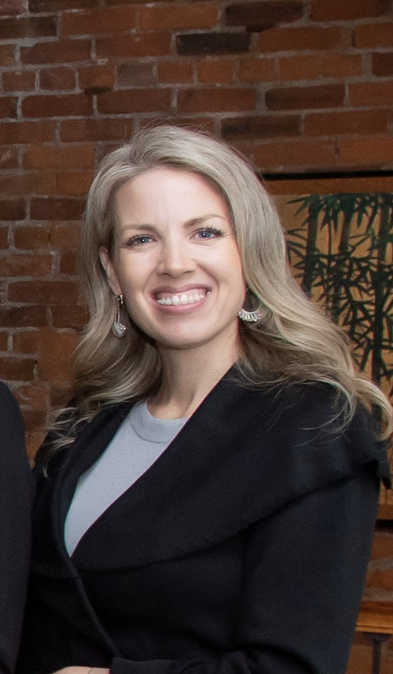 Smiling woman with blonde hair, wearing a black blazer and earrings, in front of a brick wall and artwork.