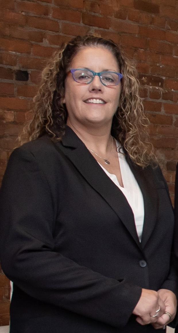 A woman with curly hair and glasses, wearing a black blazer and white blouse, standing in front of a brick wall.