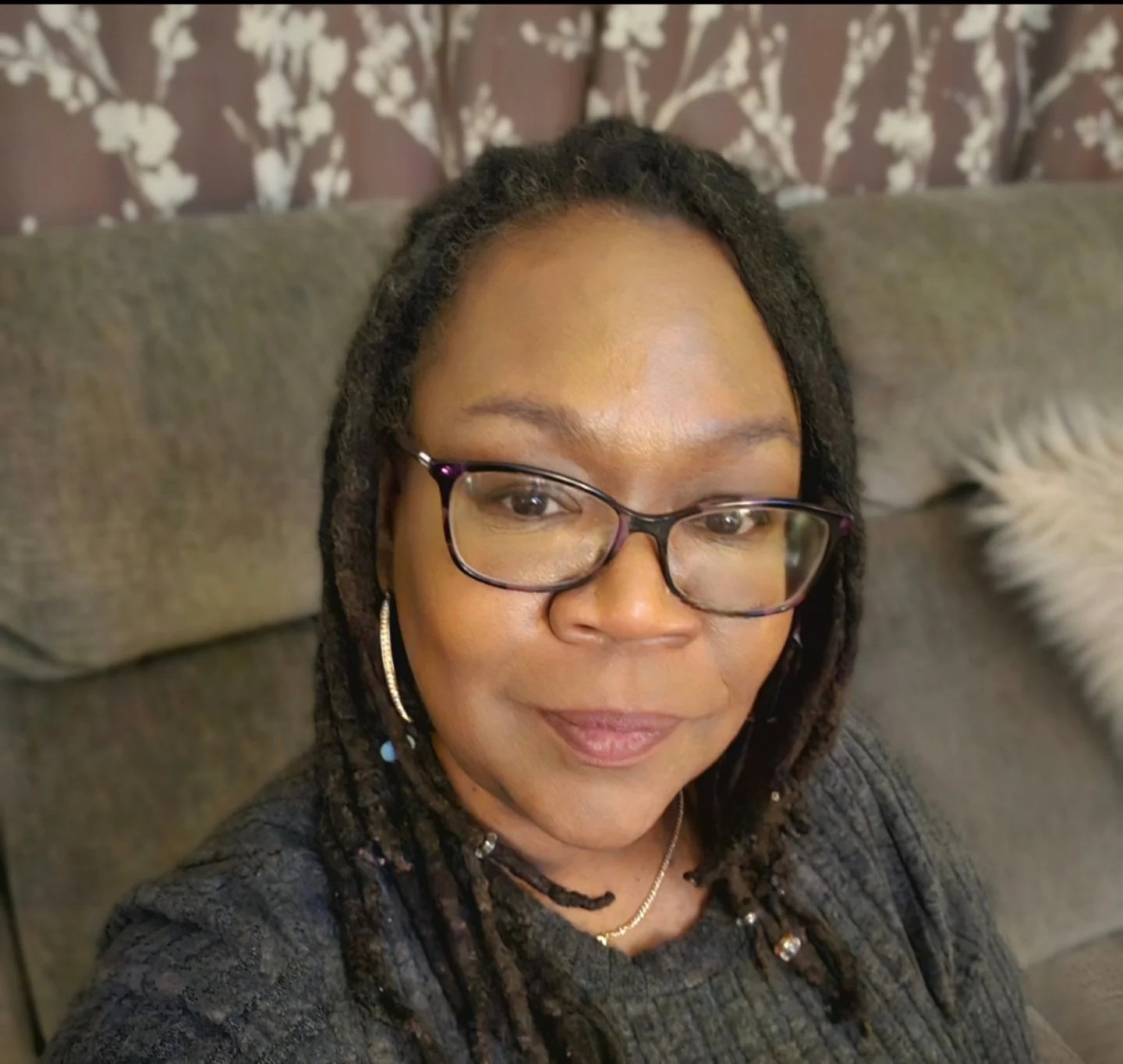 A woman with glasses and dreadlocks taking a selfie on a gray couch with a patterned curtain in the background.