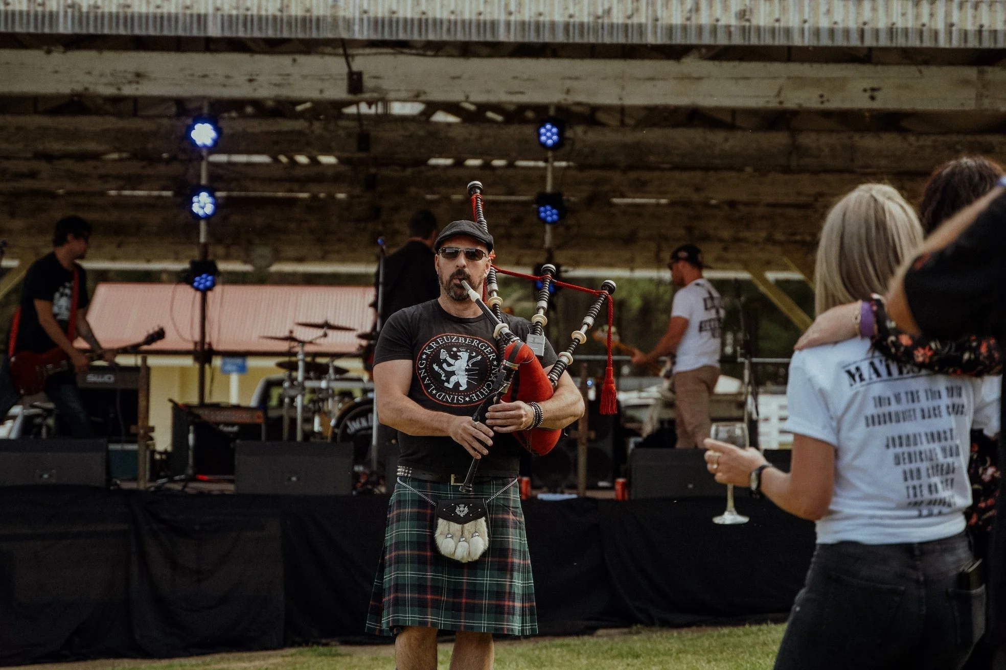 A man playing bagpipes at an outdoor music concert with a band performing on stage behind him